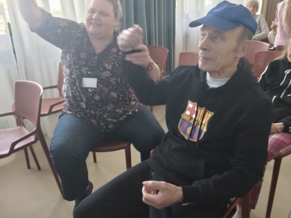 Na zdjęciu siedzący starszy mężczyzna w niebieskiej czapce trzyma za rękę siedzącą dalej kobietę ,kobieta podnosi rękę do góry w tanecznej pozie.