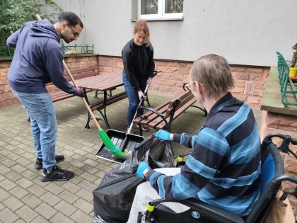 Na zdjęciu widać osoby sprzątające teren ogrodu. Mężczyzna na wózku trzyma worek na liście,wolontariusze zgarniają liście na szuflę w tle ławki ogrodowe .