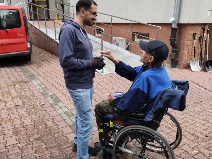 Na zdjęciu mężczyzna na wózku w niebieskiej kurtce robi gest dłonią w kierunku wolontariusza ,który uśmiecha się do niego przyjaźnie,obaj panowie prowadzą konwersację.W tle widać fragment budynku.
