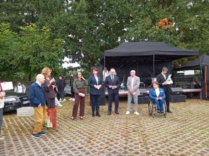 Na zdjęciu widać kobietę przemawiającą do stojących w szeregu osób obok mężczyzna w średnim wieku trzyma obraz w tle sprzęty muzyczne.