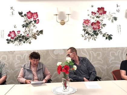Na zdjęciu grupa osób siedzi przy białym stole mężczyzna pochyla się w stronę kobiety czytającej tekst przed nimi wazon z czerwonymi kwiatami w tle piękny mural w czerwone kwiaty