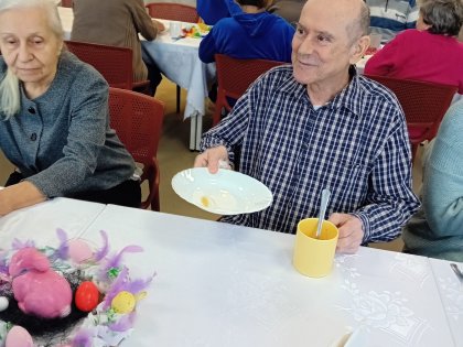 Na zdjęciu dwoje starszych ludzi spożywających posiłek, Pan z uśmiechem podaje talerz w tle inne osoby