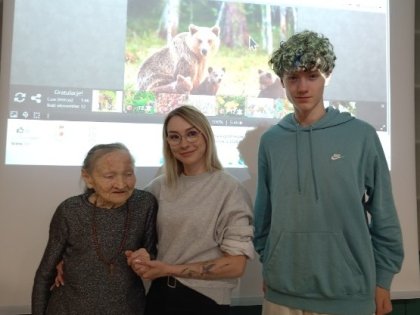 Na zdjęciu widać trójkę osób stojących pod dużym kolorowym ekranem,młody chłopak z dziewczyną i starsza pani