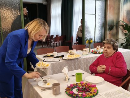 Zdjęcie przedstawia kobietę podającą posiłek starszej pani ,w tle długi stół z potrawami i wystrojem Wielkanocnym