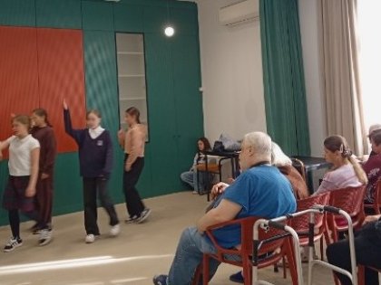 na zdjęciu grupa taneczna młodych osób jedna osoba podnosi rękę druga zwrócona w jej stronę śpiewa na widowni z przodu obrazka siedzący męszcyzna i starsza kobieta przyglądają się scenie