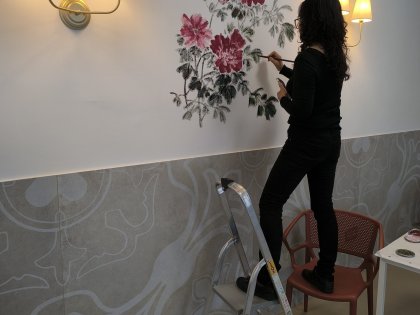 Chińska malarka stojąc na drabinie maluje pędzelkiem na białej ścianie czerwone kwiaty.