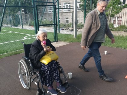 na zdjęciu po lewej widać starszą kobietę na wózku trzymającą piłeczkę do gry nieco dalej z prawej strony maszerujący w jej strone męszczyzna
