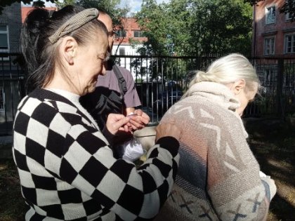 Na obrazku odwrócona starsza kobieta w beżowym swetrze za nią stoi druga kobieta w swetrze w szachownicę w tle zieleń ogrodu