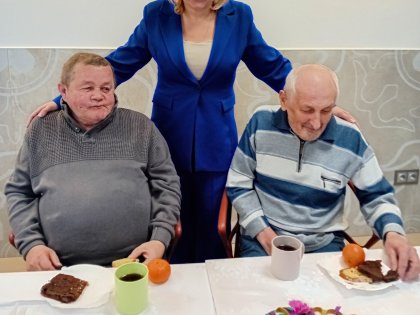 Na zdjęciu dwóch starszych panów pozuje do zdjęcia siedząc przy białym stole,z tyłu za nimi stoi kobieta uśmiecha się, ma niebieski strój trzyma panów za ramię