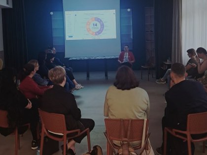 Na zdjęciu widać grupę osób siedzącą w okrąg na krzesłach,z przodu sali ekran z wyświetlanym slajdem ,kobieta siedząca z przodu w czerwonej marynarce czyta odwrócona do słuchających uczestników