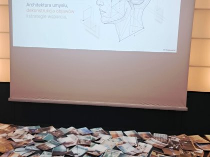 Na zdjęciu widać biały ekran z napisem zaburzenia neuropoznawcze obok rysunek ludzkiej głowy z zarysem mózgu, z przodu na długim stole porozkładane kolorowe kartki