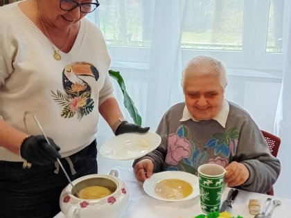 Na zdjęciu kobieta nalewa zupę starszej Pani siedzącej przy stole