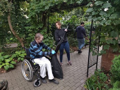 na zdjęciu mężczyzna na wózku trzyma czarny worek ogrodowy do którego wolontariuszka wrzuca szuflą liście ,w tle widać ogród i innego pracującego mężczyznę.