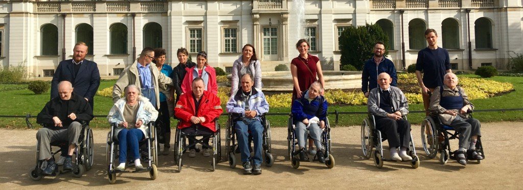 Mieszkańcy Domu na wózkach oraz pracownicy i wolontariusze pozują do wspólnego zdjęcia na tle pałacu z fontanną i klombem kwiatów.