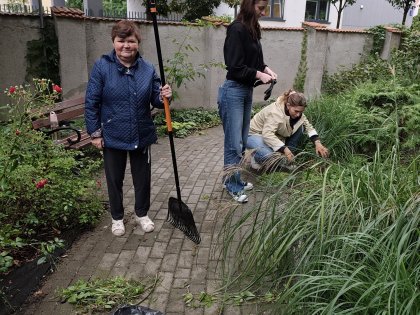 Na zdjęciu dwie panie wolontariuszki pielą rośliny, z przodu widać panią w niebieskiej kurtce trzymającą szpadel w tle piękny ogród.