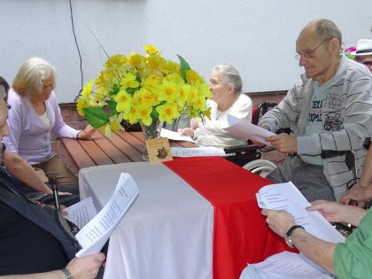 Seniorzy siedzą przy stole który ozdobiony jest Flagą Polski i żółtym bukietem kwiatów
