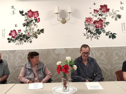 Na zdjęciu grupa osób dwóch mężczyzn i dwie kobiety siedzą przydługim białym stole przed nimi kartki z tekstem wazon z kwiatami w tle na białej ścianie piękny mural z motywem kwiatowym