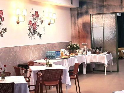 Na zdjęciu duża oświetlona sala po lewej przy białej ścianie z kolorowym muralem ustawione stoliki z białymi obrusami na stolikach widać talerzyki z ciastkami i owocami