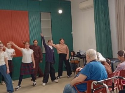 Na zdjęciu grupa młodych osób wykonuje taniec z podniesionymi rękoma przed nimi starszy mężczyzna i kobieta  przyglądając się występowi w pozycji siedzącej na czerwonych krzesłach