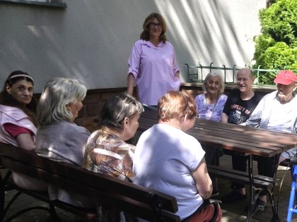 Na zdjęciu widać grupę osób siedzącą przy drewnianym stole patrzą na stojącą kobietę w różowej koszuli.Wszyscy są radośni