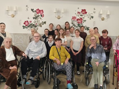 Na zdjęciu widać starsze osoby z tyłu stoi młodzież pod muralem w kolorowe kwiaty na tle białej ściany