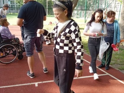 Na obrazku widać kobietę trzymającą biały plastikowy kubek ,kobieta próbuje przejść po boisku,w tle inni młodzi zawodnicy
