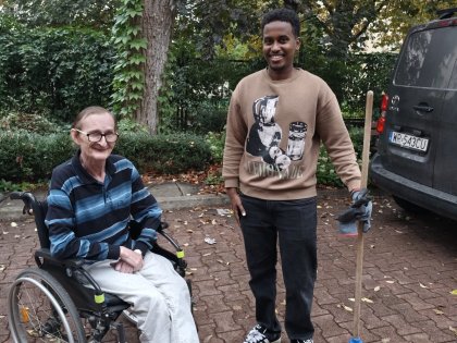 Na zdjęciu widać mężczyznę na wózku obok czarnoskóry wolontariusz trzyma miotełkę ogrodową,obaj panowie z uśmiechem pozują do zdjęcia.W tle ogród.