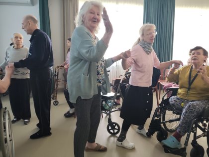 Na zdjęciu starsza Pani w atmosferze wesołej zabawy robi gest taneczny unosząc rękę, w tle  kobieta blondynka w różowym swetrze trzyma za dłonie tańczące osoby tańczące osoby.