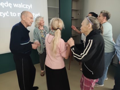 Na zdjęciu taneczny krąg starszych osób trzymających się za rękę w tle biały ekran z tekstami piosenki