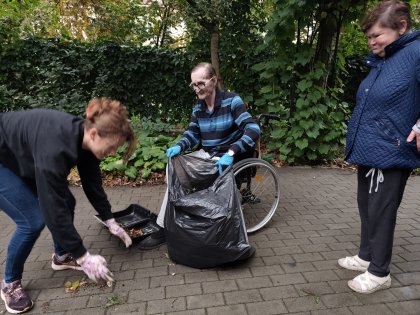 Na zdjęciu w ogrodzie wolontariuszka pochylając się sprząta liście obok niej mężczyzna na wózku trzyma czarny worek ogrodowy , z boku z zaciekawieniem przygląda się temu kobieta w niebieskiej kurtce, w tle widać zieleń ogrodu.