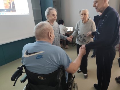N zdjęciu mężczyzna na wózku inwalidzkim trzyma starsze osoby za rękę w kręgu tanecznym
