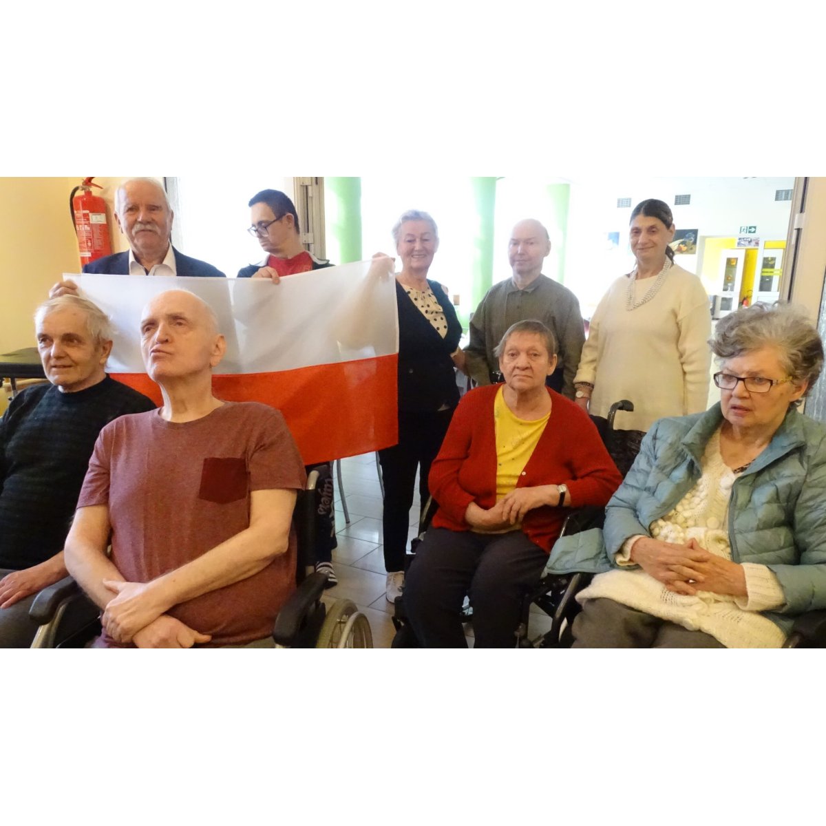 Mieszkańcy, część na wózkach, trzymają biało-czerwoną flagę podczas wspólnego śpiewania hymnu