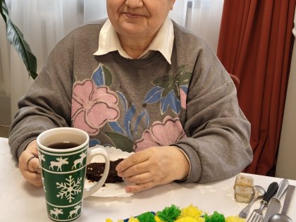 Na zdjęciu starsza pani z zadowoleniem spożywa ciasto przy udekorowanym świątecznie stole Wielkanocnym
