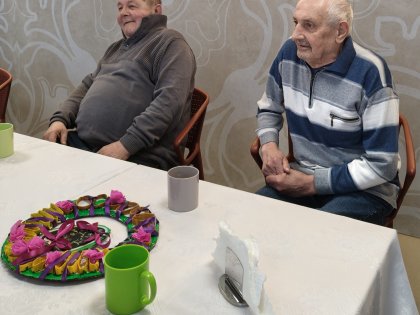Dwaj starsi panowie siedzący przy białym stole z zastawą i stroikiem Wielkanocnym, są w dobrym nasrroju