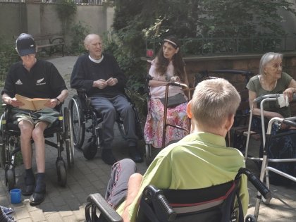 Na zdjęciu widać starsze osoby siedzące przy drewnianym stole w radosnych pozach w tle ogród