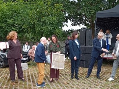 Na zdjęciu widać gromadę osób w wesołym piknikowym nastroju,w centrum stoi mężczyzna z kobietą ,rozmawiają ze sobą,kobieta trzyma obraz.