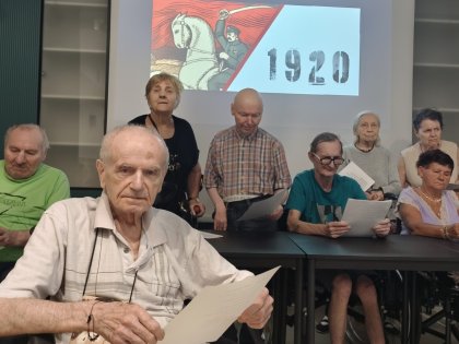 Na zdjęciu starszy mężczyzna czyta wiersz w tle inne osoby przy stole z kartkami wierszy