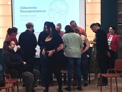 Na zdjęciu grupa ludzi rozproszona na sali z przodu jasny ekran z widocznym slajdem na slajdzie napis zaburzenia neuropoznawcze obok rysunek ludzkiej głowy z zarysem mózgu.