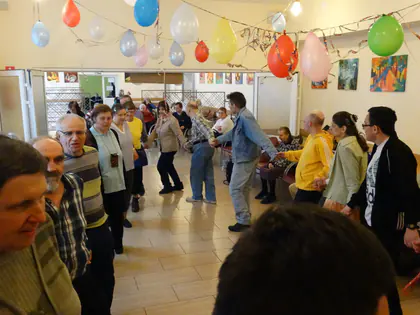 Uczestnicy zabawy tańczą w kole w sali udekorowanej balonami i kolorowymi obrazami