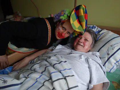 Klaun w kolorowej czapce pochyla się nad uśmiechniętą mieszkanką leżącą w łóżku