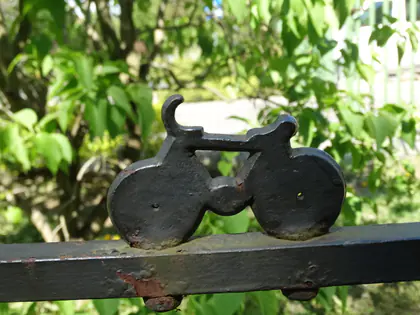 Metalowa ozdoba w kształcie roweru na ogrodzeniu, w tle zielone liście