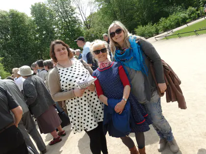 Trzy uśmiechnięte kobiety pozują w parku, jedna w niebieskiej chuście i okularach