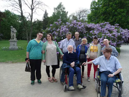 Grupa mieszkańców, część na wózkach, przy kwitnących rododendronach i rzeźbie w Łazienkach