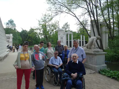 Mieszkańcy, część na wózkach, stoją w parku Łazienek Królewskich przed rzeźbą i kolumnadą