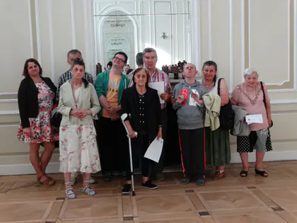 Mieszkańcy i opiekunowie pozują w jasnej sali, część osób trzyma kartki