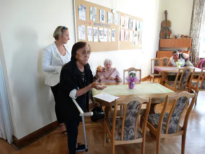 Kobieta o kuli rozmawia z pracowniczką w jasnym żakiecie w jadalni, w tle stoły z kwiatami i tablica ze zdjęciami