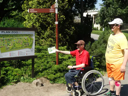 Mieszkaniec na wózku wyciąga rękę z folderem przy tablicy z planem ZOO i drogowskazem, obok stoi opiekun w czapce