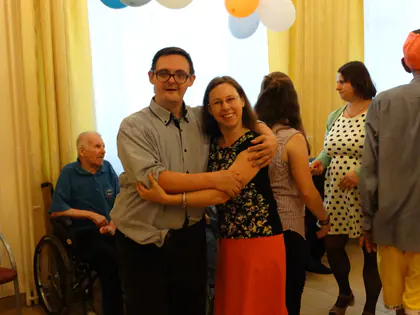 Mieszkaniec w okularach obejmuje wolontariuszkę podczas zabawy, obok starszy mężczyzna na wózku
