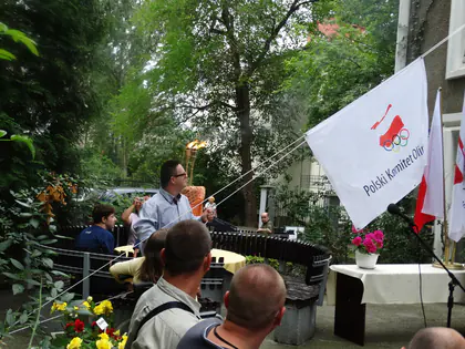 Biała flaga Polskiego Komitetu Olimpijskiego i flagi Polski w ogrodzie, w tle mówca przy stole