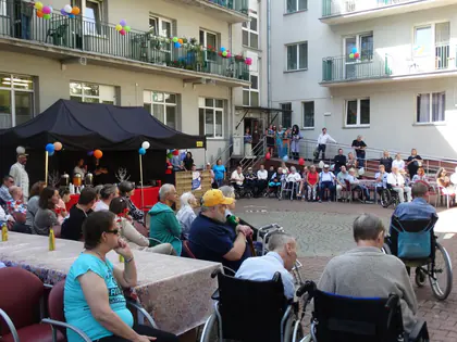 Mieszkańcy siedzą na dziedzińcu Domu pod namiotem, na balkonach wiszą balony